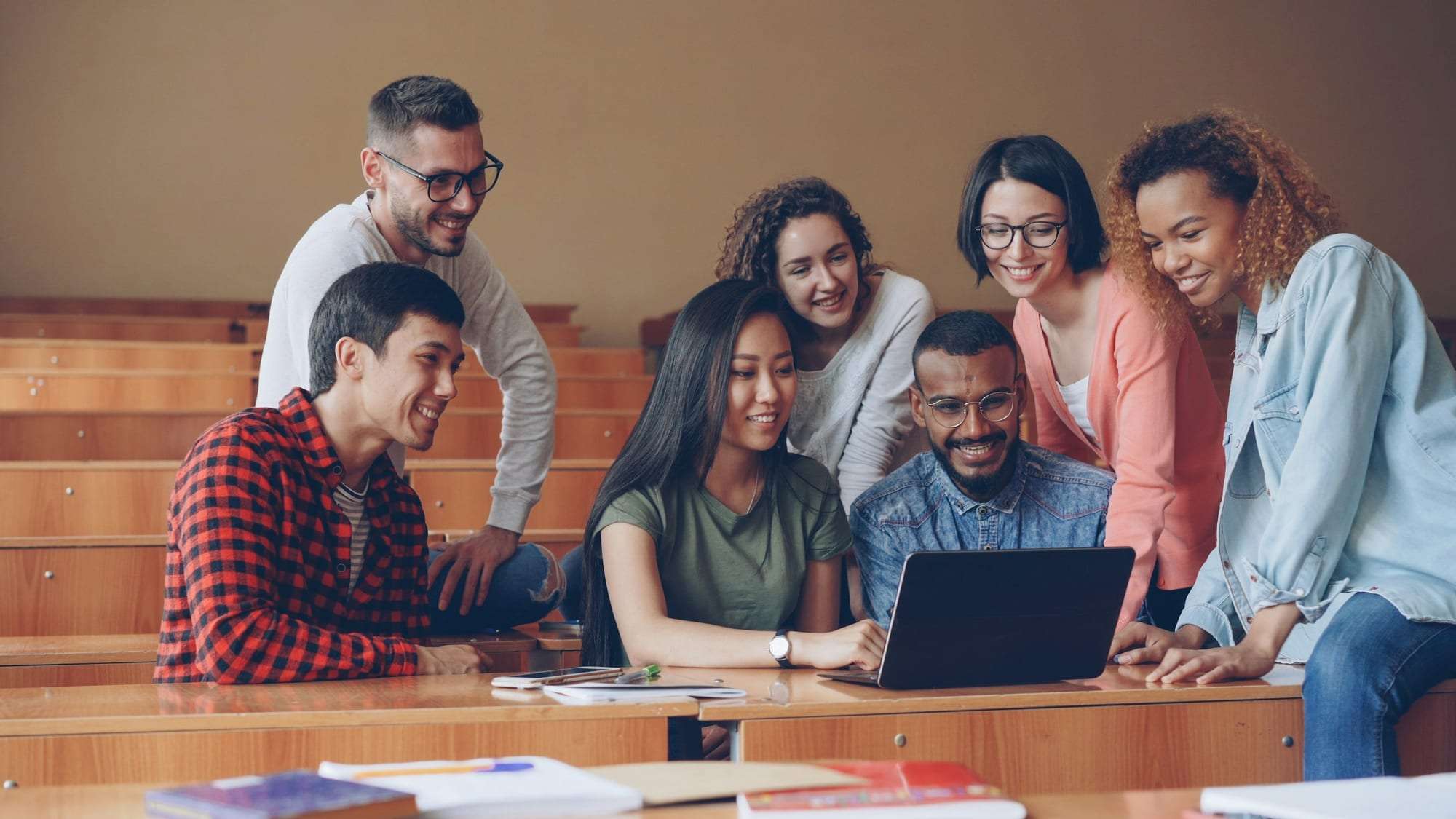 A mixed group of students gathered around a laptop in a lecture hall, smiling