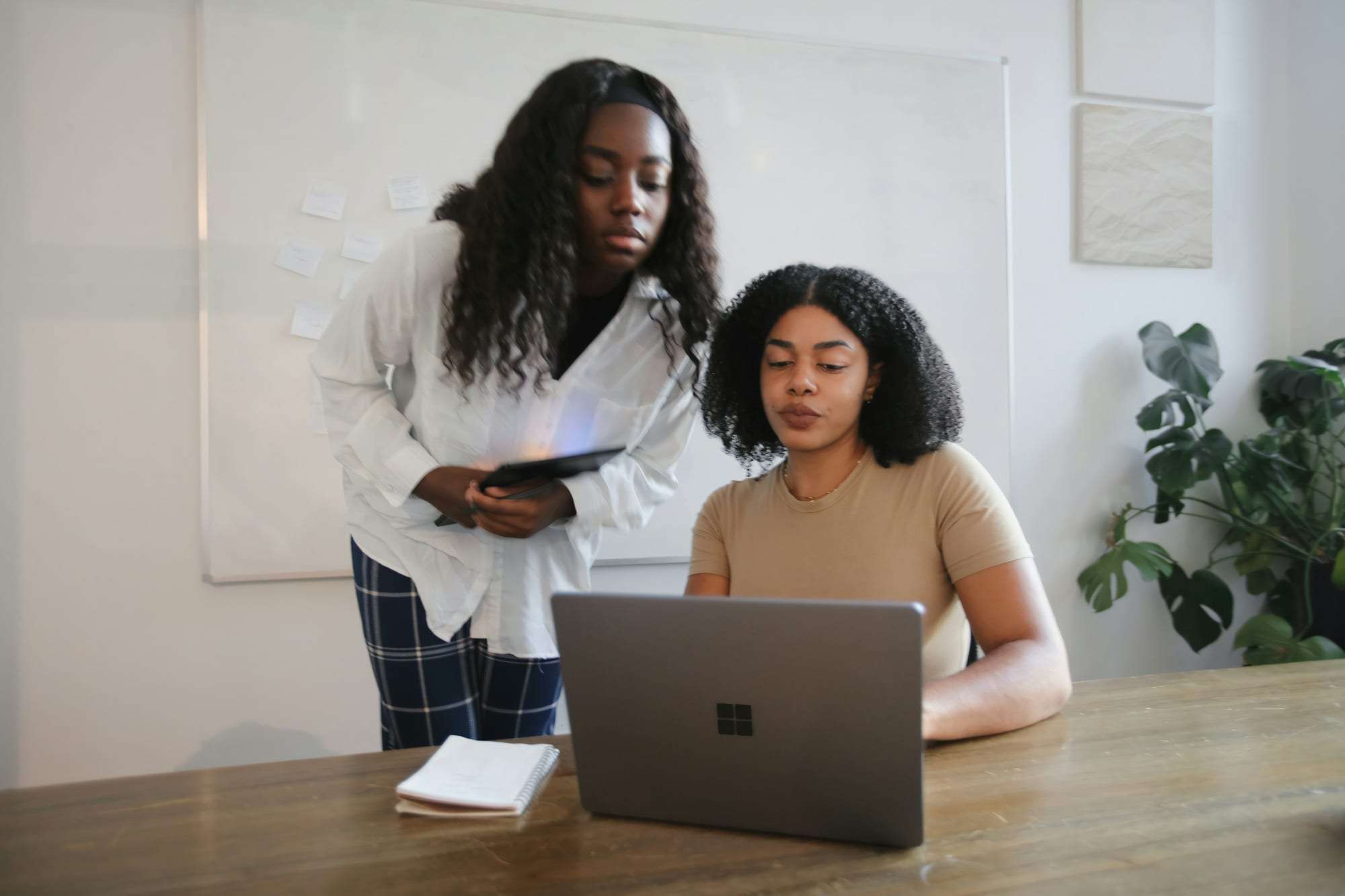 Two young women at a laptop, one standing with a tablet and one seated, reading the screen together