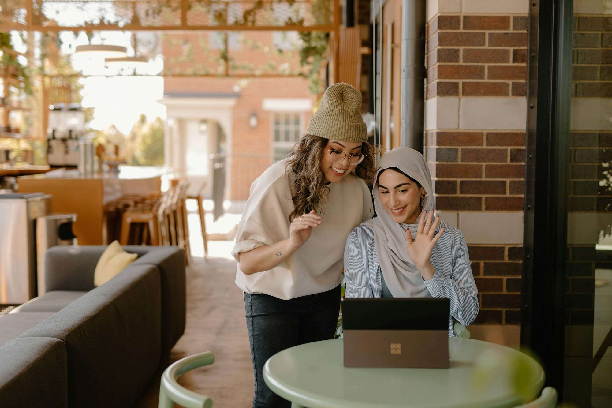 Two young women smiling at a laptop in a café, one in a hijab waving at the screen during a video call