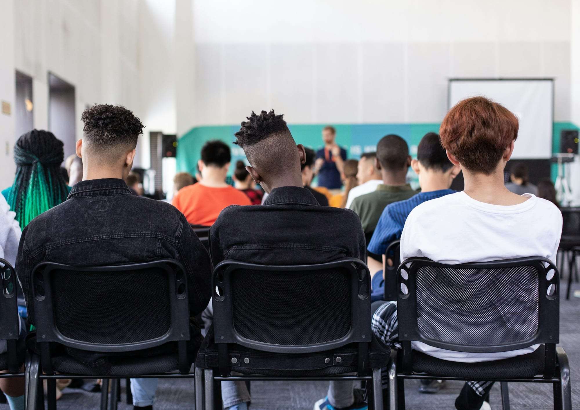 Young people from different backgrounds seated in rows, seen from behind, listening to a speaker at the front of a room