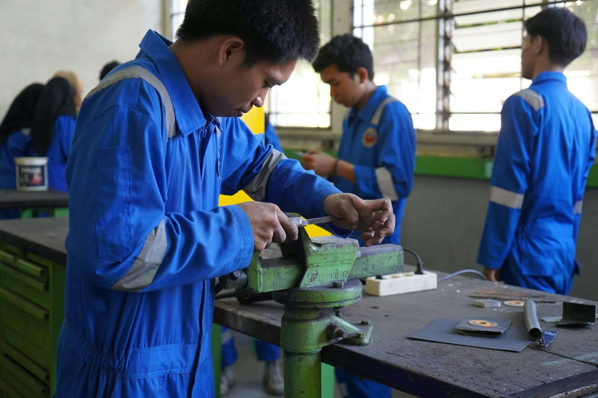 Trainees in blue overalls working at bench vices in a technical workshop
